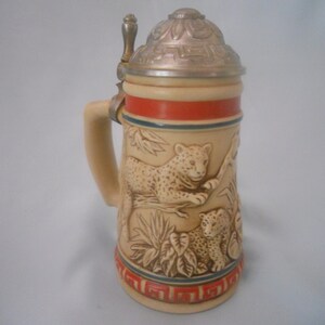 May include: A ceramic beer stein with a lid featuring a detailed design of two leopards in a jungle setting. The stein has a red and blue band around the middle.
