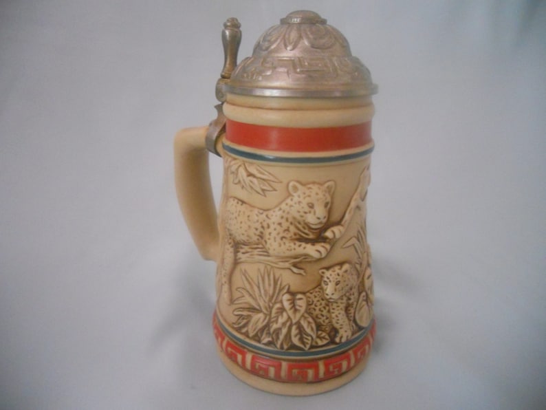 May include: A ceramic beer stein with a lid featuring a detailed design of two leopards in a jungle setting. The stein has a red and blue band around the middle.
