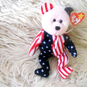 May include: A plush teddy bear with a patriotic design. The bear features a white face, red and white striped arms and legs, and a navy blue body with white stars. A red heart-shaped tag is attached.