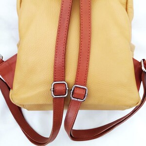May include: A yellow leather backpack with brown leather straps and accents. The backpack has two adjustable shoulder straps with silver-colored metal buckles. The bag's design includes a top handle and a flap closure.