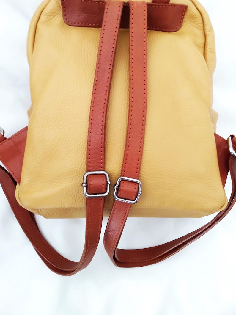 May include: A yellow leather backpack with brown leather straps and accents. The backpack has two adjustable shoulder straps with silver-colored metal buckles. The bag's design includes a top handle and a flap closure.
