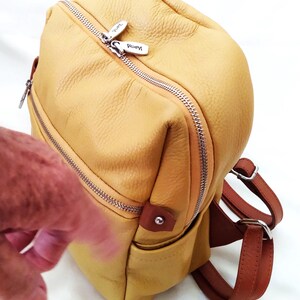 May include: A yellow leather backpack with brown straps and silver zippers. The bag has a rounded shape and a small pocket on the front. The zipper pulls are silver and have the brand name "Vemmy" on them.