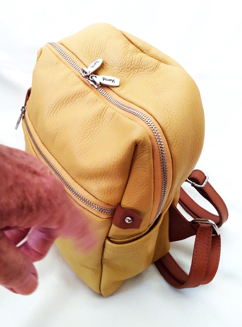 May include: A yellow leather backpack with brown straps and silver zippers. The bag has a rounded shape and a small pocket on the front. The zipper pulls are silver and have the brand name "Vemmy" on them.