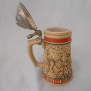 May include: A ceramic beer stein with a pewter lid features a detailed relief of a leopard in a jungle setting. The stein has a tan base with a red and blue stripe around the top.