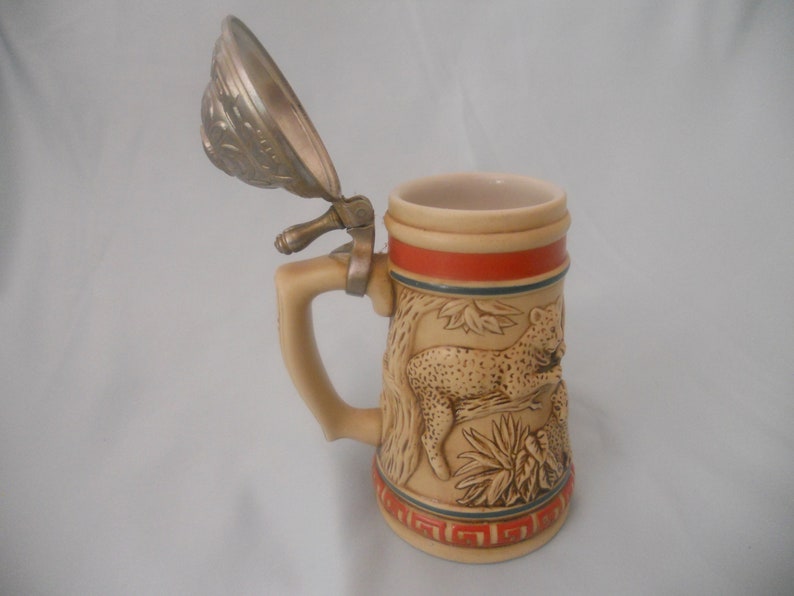 May include: A ceramic beer stein with a pewter lid features a detailed relief of a leopard in a jungle setting. The stein has a tan base with a red and blue stripe around the top.