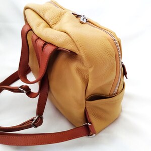 May include: A tan leather backpack with brown straps and silver-tone hardware. The backpack has a zippered closure and a small pocket on the side. The leather has a textured appearance.