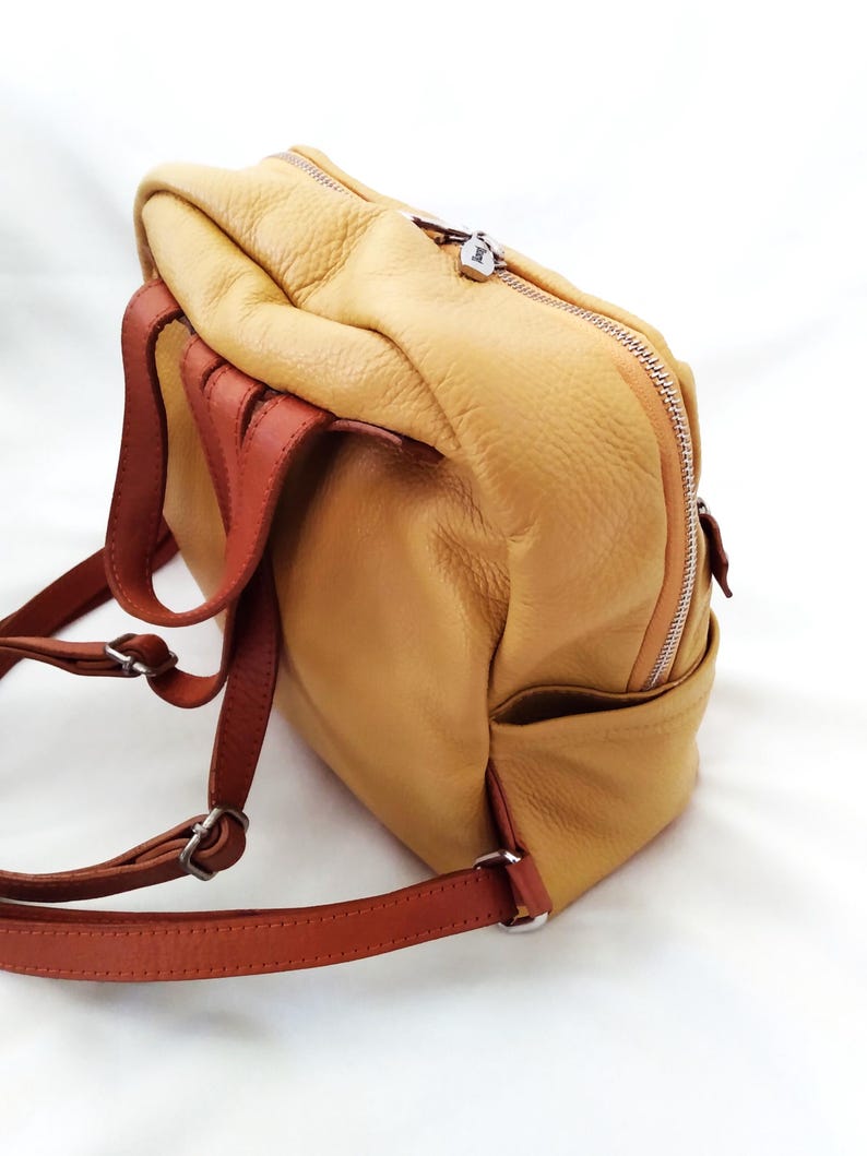 May include: A tan leather backpack with brown straps and silver-tone hardware. The backpack has a zippered closure and a small pocket on the side. The leather has a textured appearance.