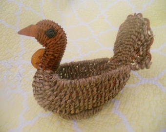May include: A woven wicker basket shaped like a turkey. The basket is brown and has a natural finish. It has a small opening at the top for holding items.