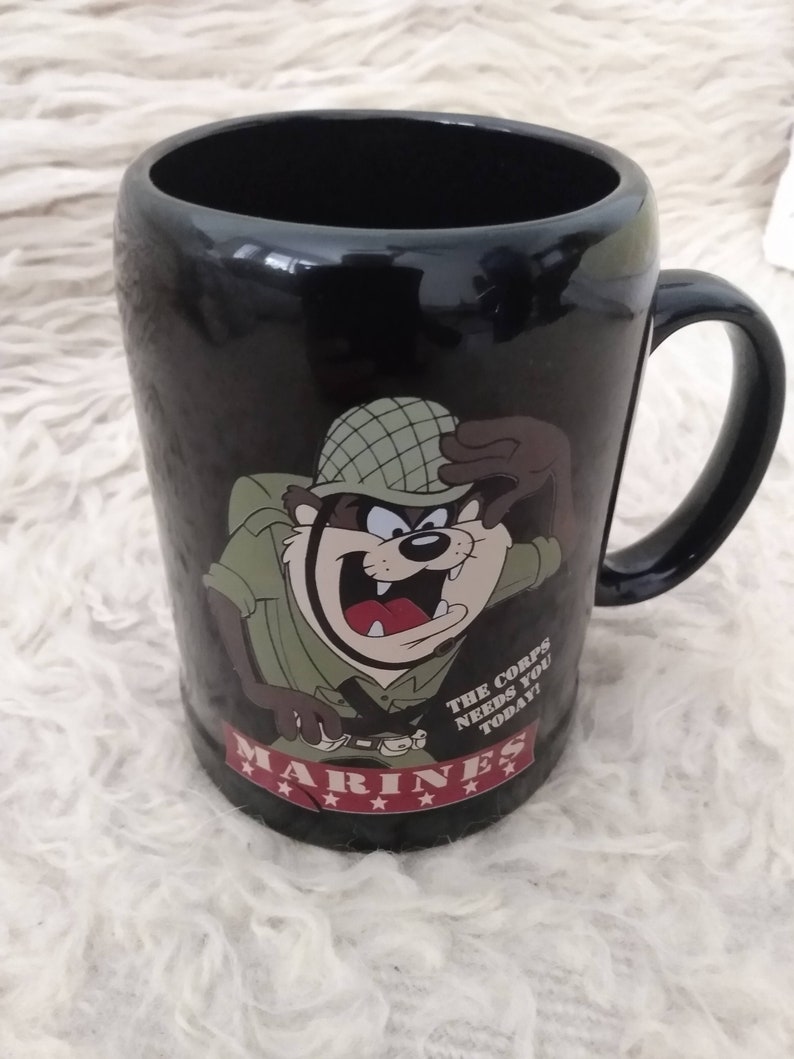 May include: Black ceramic mug featuring a cartoon image of a Tasmanian Devil dressed as a Marine. The mug has the text "The Corps Needs You Today!" and "Marines" with five stars below the text.