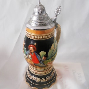 Może przedstawiać: Ceramiczny kufel do piwa z czarnym tłem i kolorową sceną ludzi pijących piwo. Kufel ma srebrną pokrywkę z uchwytem na kciuk i uchwytem. Podstawa kufla jest ozdobiona wzorami kwiatowymi.