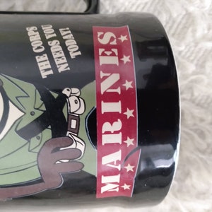 May include: Black ceramic mug featuring a cartoon character in a green military uniform with the text "The Corps Needs You Today!" and "MARINES" with five red stars.