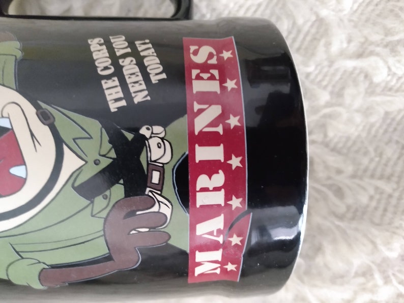 May include: Black ceramic mug featuring a cartoon character in a green military uniform with the text "The Corps Needs You Today!" and "MARINES" with five red stars.