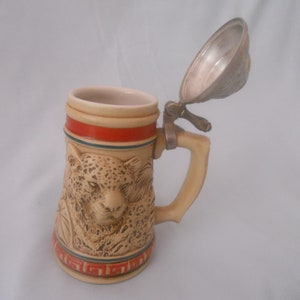 May include: A ceramic beer stein with a hinged lid features a detailed relief of a leopard's face. The stein has a brown and beige base with red, blue, and white accents.