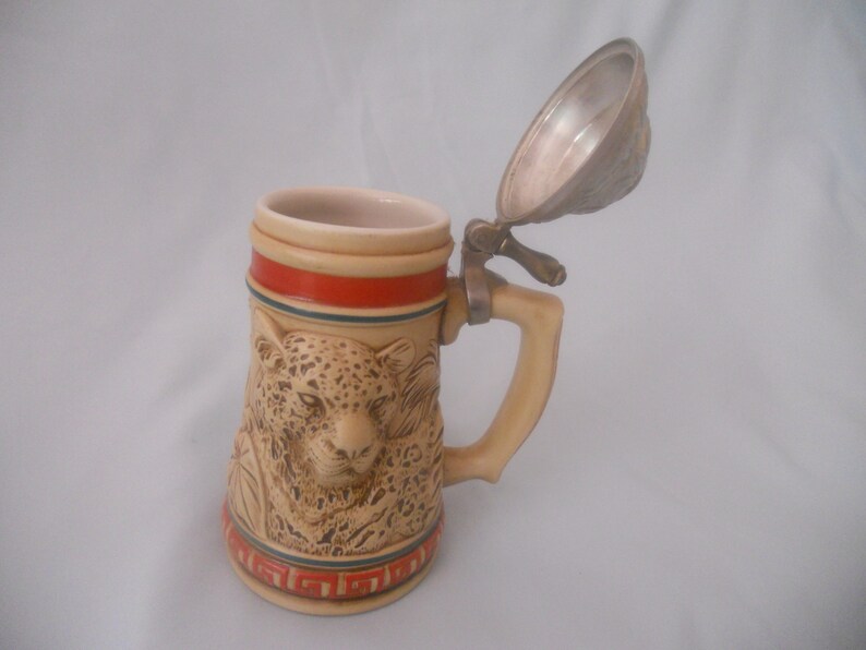 May include: A ceramic beer stein with a hinged lid features a detailed relief of a leopard's face. The stein has a brown and beige base with red, blue, and white accents.