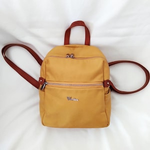 May include: Mustard yellow backpack with brown straps and a top handle. The bag has a zippered main compartment and a smaller front zippered pocket. A small silver logo is visible on the front.