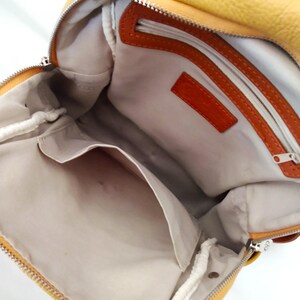 May include: Open view of a yellow leather backpack with a silver zipper. The interior features a light-colored lining, a small pocket, and orange leather accents. The bag is designed with a single strap.