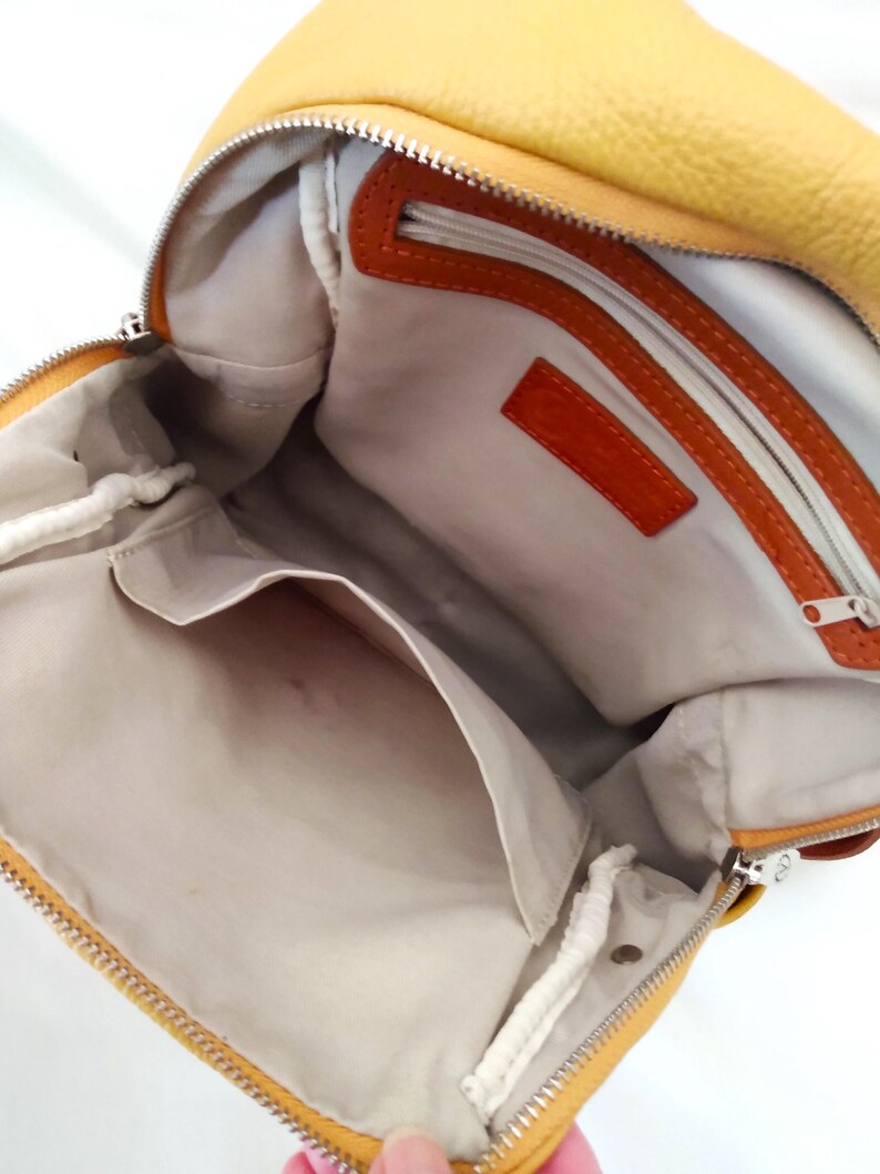 May include: Open view of a yellow leather backpack with a silver zipper. The interior features a light-colored lining, a small pocket, and orange leather accents. The bag is designed with a single strap.