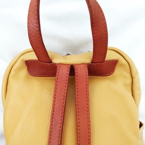 May include: A yellow leather backpack with brown leather straps and handle. The backpack has a rounded shape and a textured surface. The straps are stitched and appear durable. The bag is designed for carrying items.