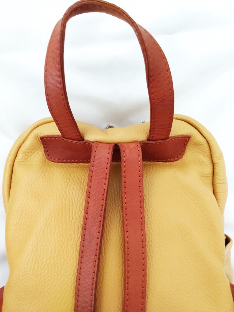May include: A yellow leather backpack with brown leather straps and handle. The backpack has a rounded shape and a textured surface. The straps are stitched and appear durable. The bag is designed for carrying items.