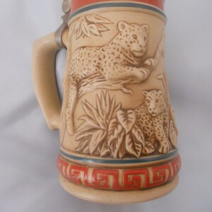 May include: A ceramic beer stein with a brown and white design of two leopards in a jungle setting. The stein has a metal lid and a handle.