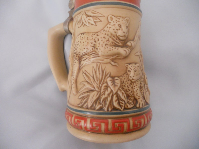 May include: A ceramic beer stein with a brown and white design of two leopards in a jungle setting. The stein has a metal lid and a handle.