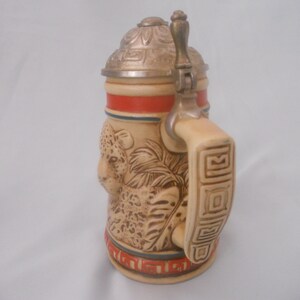May include: A ceramic beer stein with a lid and handle. The stein features a detailed image of a leopard with a jungle background. The lid is silver with a decorative design. The handle is decorated with a geometric pattern.