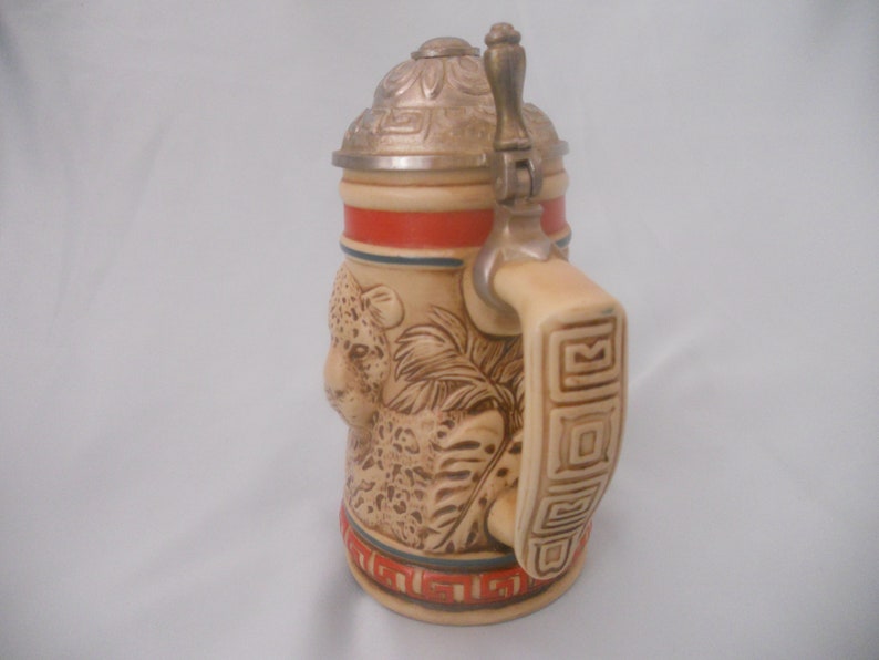 May include: A ceramic beer stein with a lid and handle. The stein features a detailed image of a leopard with a jungle background. The lid is silver with a decorative design. The handle is decorated with a geometric pattern.