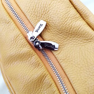 May include: Close-up of a mustard yellow leather bag with a silver zipper. The zipper pulls are silver with the word "Valentina" engraved on them. The leather has a textured appearance.
