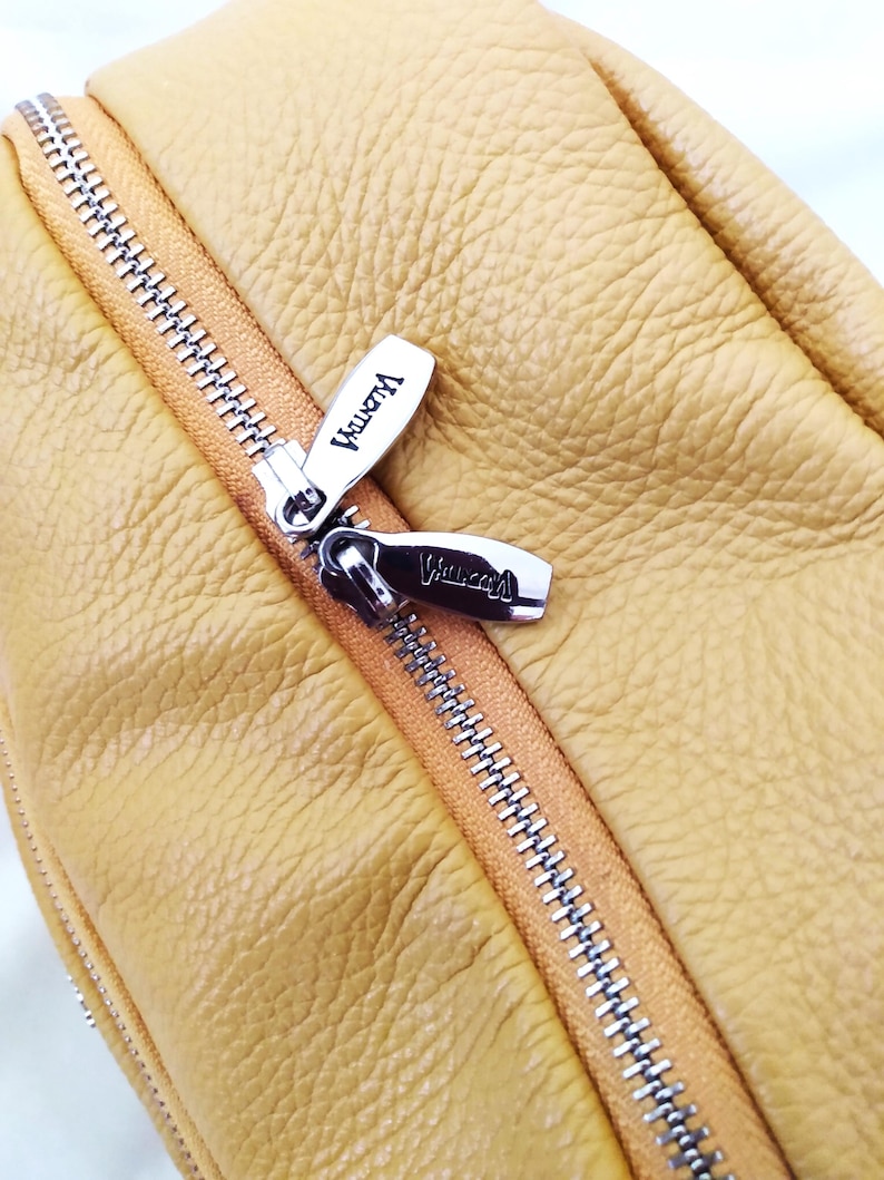 May include: Close-up of a mustard yellow leather bag with a silver zipper. The zipper pulls are silver with the word "Valentina" engraved on them. The leather has a textured appearance.