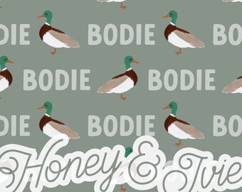 May include: A repeating pattern of illustrated ducks with green heads, brown bodies, and white chests on a gray background. The word "BODIE" is interspersed between the ducks. The text "Honey & Tvie" is also present.