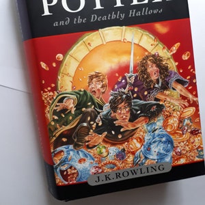 May include: A black hardback book with the title "Harry Potter and the Deathly Hallows" in white text. The cover features an illustration of three characters, Harry Potter, Ron Weasley, and Hermione Granger, surrounded by gold coins and other magical objects.