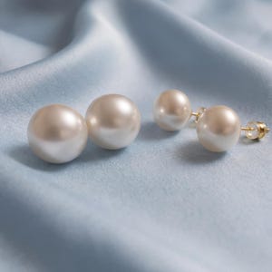 May include: A pair of stud earrings featuring large, round, lustrous white pearls. Each earring is crafted with a gold-tone metal post and a secure backing, showcasing a smooth, reflective surface that catches the light with a soft glow.