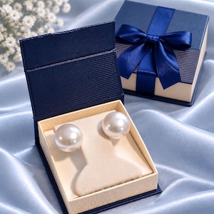 May include: Two large, round, white pearl stud earrings are displayed on a beige velvet insert inside an open, dark blue, textured square jewelry box. A matching closed dark blue box with a blue satin ribbon bow sits in the background.