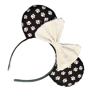 May include: A headband featuring two round ears covered in black fabric with a white paw print pattern. A large, white, sequined bow is attached to the center between the ears. The headband frame is thin, black, and curved to fit a head.