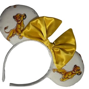 May include: White headband with Simba-themed ears and a large gold satin bow. The ears feature a Simba cartoon print on a white background. The headband is silver. A fun accessory for fans of the Lion King.