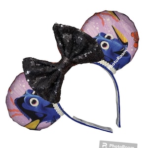 May include: A pair of black sequined Minnie Mouse ears with a bow. The ears are decorated with a purple and blue fabric featuring Dory from Finding Nemo. The text "PhotoRoom" is visible on the right ear.