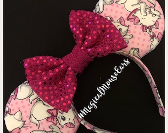 Peut inclure: Des oreilles de Minnie Mouse roses avec un tissu à pois rose et blanc représentant un chat blanc avec des accents roses. Le nœud est rose pailleté avec un centre en velours rose. Le serre-tête est blanc avec le texte #MagicalMouseEars.