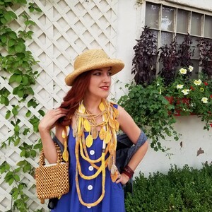 May include: A woman wearing a blue dress and a straw hat with a yellow and white crocheted leaf necklace. She is holding a wooden beaded bag.