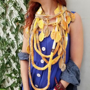 May include: A long, multi-strand crocheted scarf in shades of yellow and white. The scarf is made of chunky yarn and features a leaf pattern. The scarf is worn around the neck of a person wearing a blue dress.