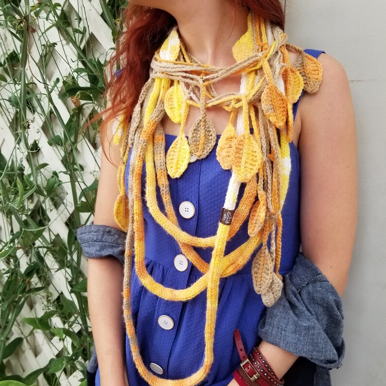 May include: A long, multi-strand crocheted scarf in shades of yellow and white. The scarf is made of chunky yarn and features a leaf pattern. The scarf is worn around the neck of a person wearing a blue dress.