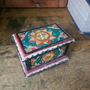 Könnte beinhalten: Eine kleine Holzkiste mit einem bemalten Design, das eine Sonne mit einem Gesicht, Sterne und Blumen zeigt. Die Kiste ist in Rot-, Grün-, Gelb- und Schwarztönen bemalt.