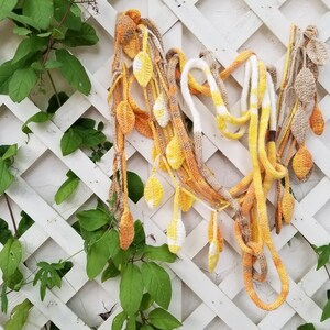 May include: A crocheted garland with yellow, orange, and brown leaves hanging on a white lattice fence. The garland is made of yarn and is a decorative item for a home or garden.