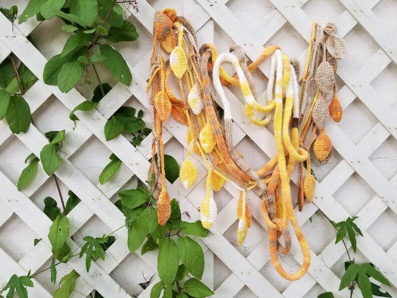 May include: A crocheted garland with yellow, orange, and brown leaves hanging on a white lattice fence. The garland is made of yarn and is a decorative item for a home or garden.