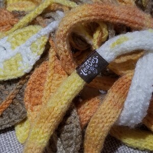 May include: A multi-coloured knitted scarf with a brown tag that says "Leaves". The scarf is made of a soft, chunky yarn and features a variety of colours, including yellow, orange, brown, and white.