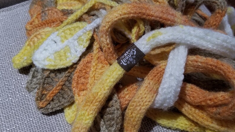 May include: A multi-coloured knitted scarf with a brown tag that says "Leaves". The scarf is made of a soft, chunky yarn and features a variety of colours, including yellow, orange, brown, and white.