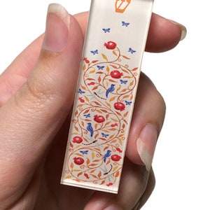 Puede incluir: Una mezuz&aacute; blanca con una estrella de David dorada en la parte superior. La mezuz&aacute; est&aacute; decorada con un dise&ntilde;o floral rojo y naranja con p&aacute;jaros azules y mariposas.