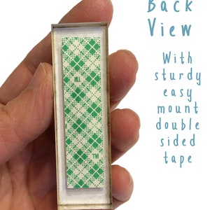 Puede incluir: Vista posterior de un letrero rectangular con un patr&oacute;n de cuadros verdes y blancos en la parte posterior. El letrero tiene un marco de pl&aacute;stico transparente y est&aacute; unido a una superficie blanca con cinta adhesiva de doble cara. La cinta est&aacute; etiquetada con "TM".