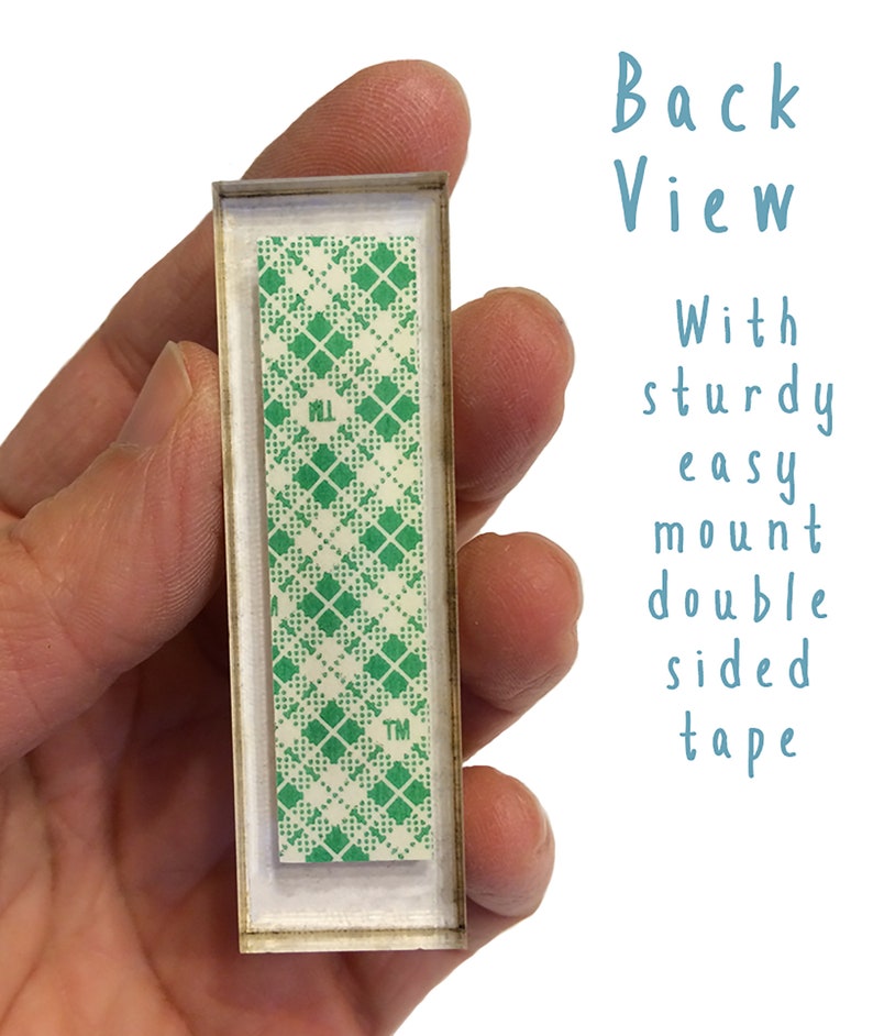 Puede incluir: Vista posterior de un letrero rectangular con un patr&oacute;n de cuadros verdes y blancos en la parte posterior. El letrero tiene un marco de pl&aacute;stico transparente y est&aacute; unido a una superficie blanca con cinta adhesiva de doble cara. La cinta est&aacute; etiquetada con "TM".