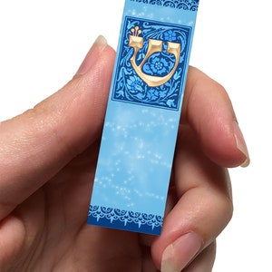 Peut inclure: Un marque-page bleu avec une lettre h&eacute;bra&iuml;que dor&eacute;e 'Heh' dans un motif floral d&eacute;coratif. La lettre est entour&eacute;e de fleurs et de feuilles bleues et blanches. Le marque-page a un fond bleu clair avec un effet de scintillement subtil.