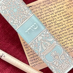 Op de afbeelding: Een blauwe en witte mezuzah met ingewikkelde bloemmotieven en het Hebreeuwse woord "Shaddai" erop. De mezuzah ligt op een stuk perkament met Hebreeuwse tekst.